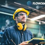 Werker mit gelbem Helm notiert Informationen auf einem Tablett in einer Produktionshalle