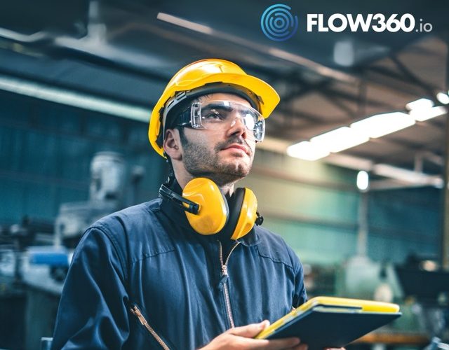 Werker mit gelbem Helm notiert Informationen auf einem Tablett in einer Produktionshalle