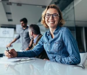 Lächelnde junge blonde Frau mit Brille und Jeanshemd, die in einem Besprechungsraum am Tisch sitzt und einen Stift hält. Im Hintergrund sind undeutlich weitere Kollegen zu sehen.